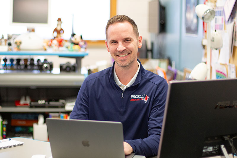 A man sits at a desk with a laptop, smiling at the camera, in a school administration office.